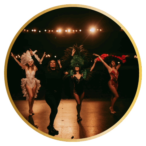 Four performers in elaborate showgirl costumes with feathered headdresses and sequined outfits dancing on stage with their arms raised. The circular-cropped photo shows them in white, green, and red costumes against a dark stage backdrop with stage lights visible in the background.
