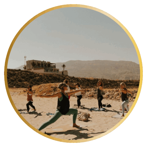 A group of people practicing yoga outdoors on yoga mats in a desert landscape, with rocky terrain, mountains in the background, and a modern building visible on a hillside. The participants are in various yoga poses on what appears to be sandy or dusty ground scattered with rocks.
